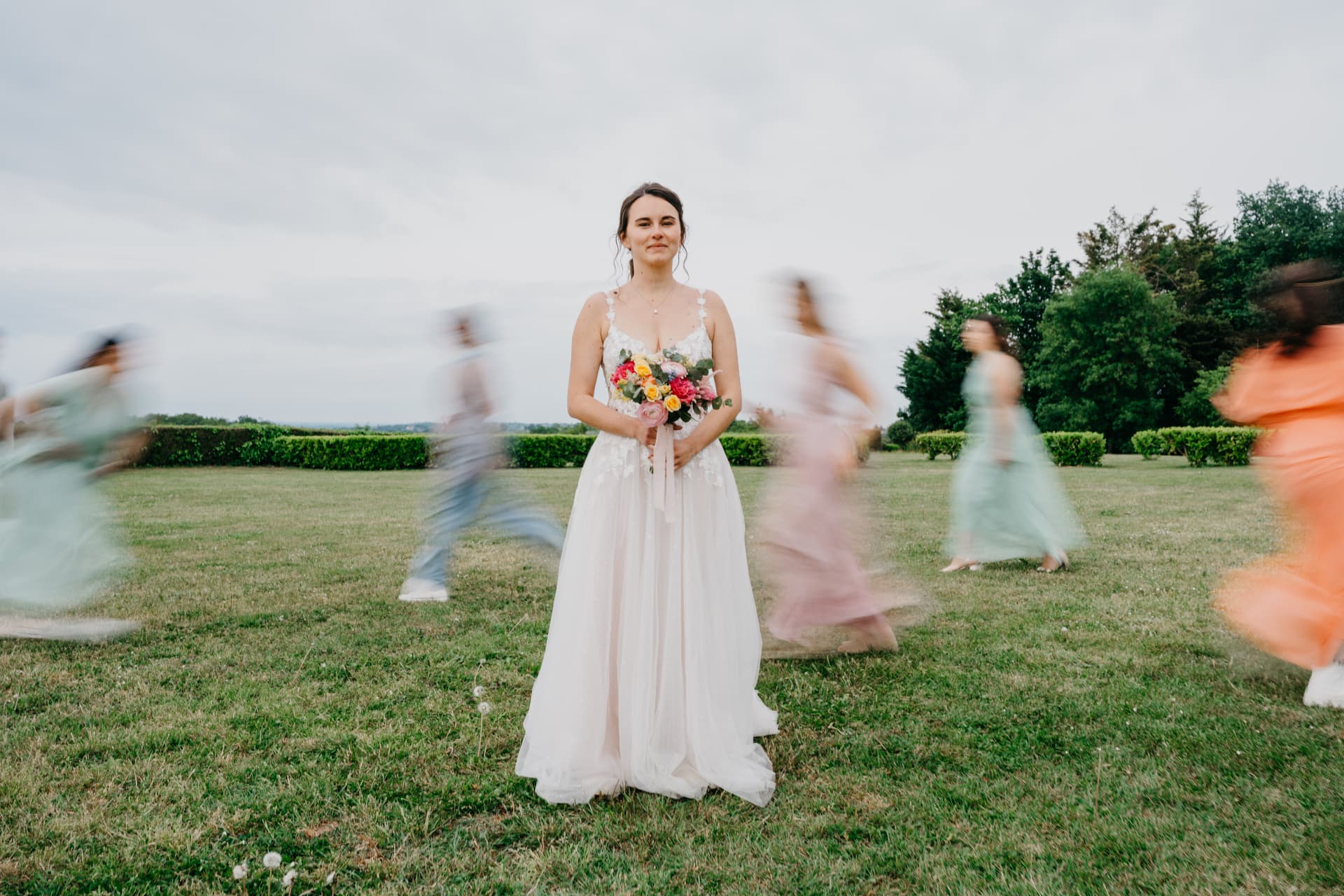 La mariée debout dans un pré, bouquet en main, entourée d'invités en mouvement