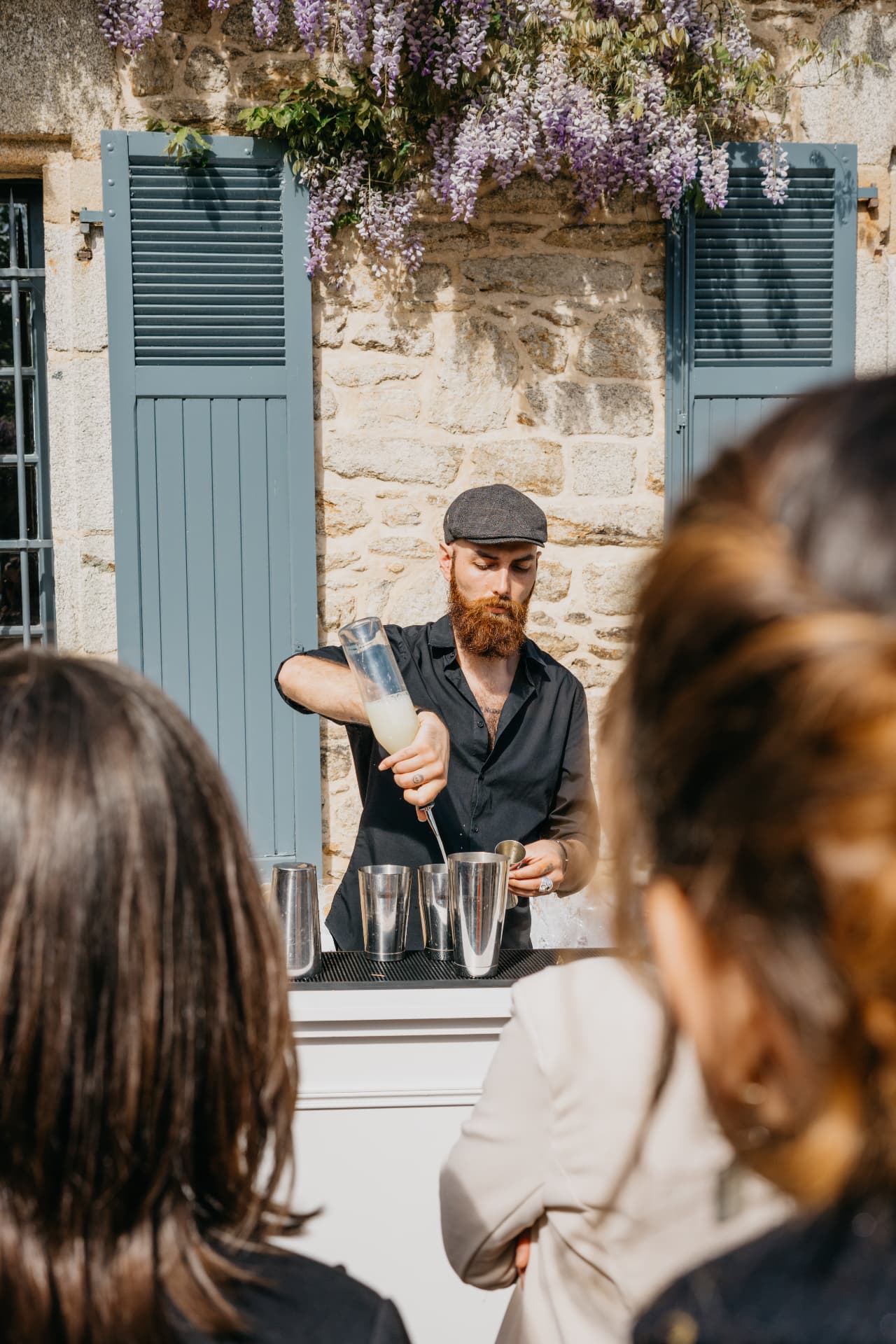 Barman au chapeau préparant un cocktail devant un mur de pierre fleuri de glycines
